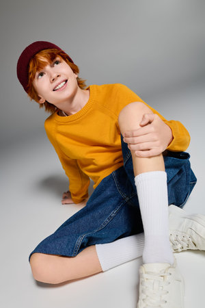 Cheerful redhead boy dressed in casual outfit sits playfully in a vibrant studio space.