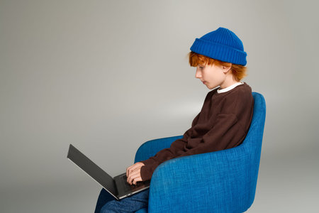Redhead boy wearing a cozy outfit sits on a chair, deeply engaged with his laptop in a studio.