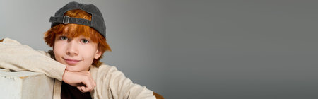 Redhead boy smiles confidently while wearing stylish casual clothes in a grey studio, banner