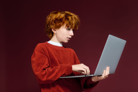 Boy with red hair uses a laptop, highlighting modern children's fashion in a studio.