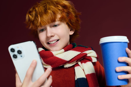 Young boy with vibrant red hair engages with his phone while holding a drink.の写真素材