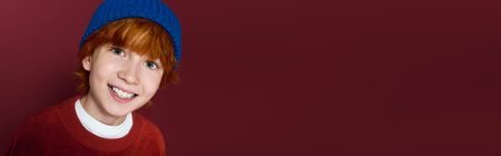 Cheerful redhead boy smiles brightly while posing in a trendy red studio, banner
