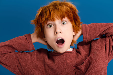 A young redhead boy expresses excitement while posing in a blue studio environment.
