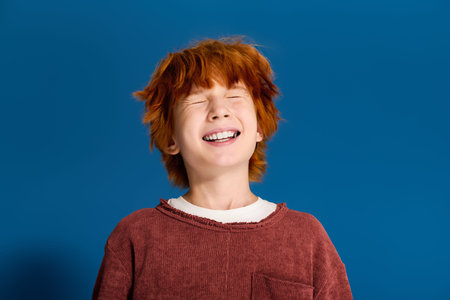 Redhead boy smiles with full energy in casual attire against a vibrant blue backdrop.の写真素材