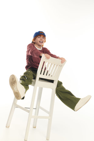 Smiling boy with red hair and casual clothes plays cheerfully on a tall chair in a studio.