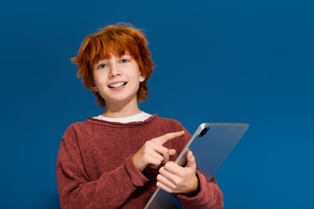 Redhead boy in casual outfit playfully interacts with his tablet against a blue backdrop.