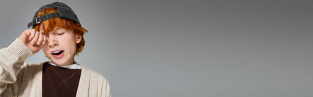 Young boy with red hair in stylish casual wear yawning in a grey studio, banner