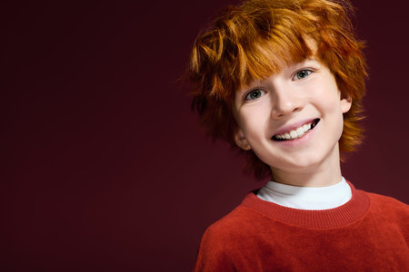 A cheerful redhead boy in casual clothes beams with joy while posing in a red studio.の写真素材
