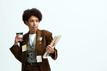 Stylish young woman carries documents and coffee against a bright backdrop.