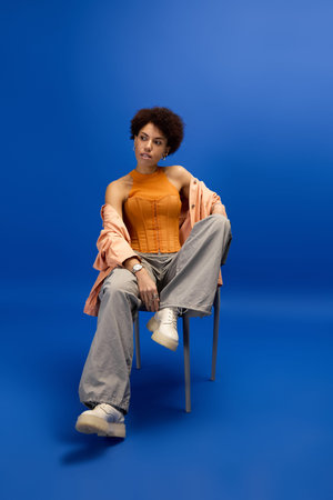 Young woman in an orange top and relaxed pants confidently poses on a chair against a blue backdrop.