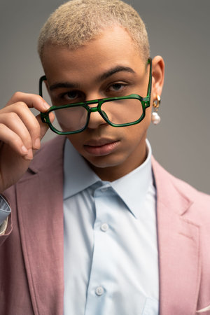 A handsome man in a pastel suit adjusts his bold glasses stylishly in a studio.