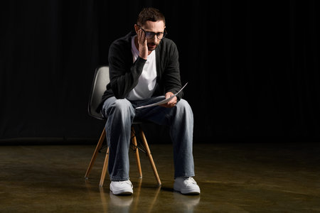 A handsome man sits on a chair, reviewing his script with deep concentration in a theater setting.