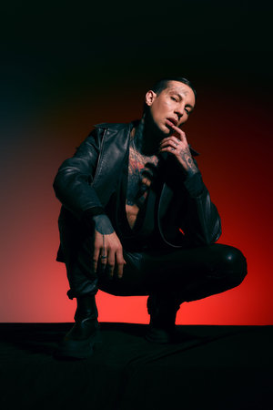 A stylish man in a leather jacket crouches confidently against a vibrant backdrop.