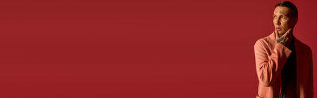 A young adult man stands thoughtfully, showcasing his modern style against a vibrant red backdrop.の写真素材