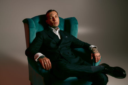 Handsome man in a tailored suit sits confidently in a teal armchair, showcasing modern fashion.