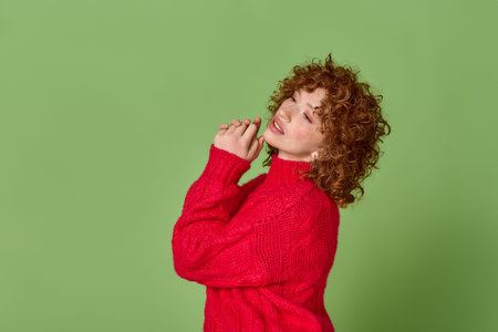 A joyful woman with red curls showcases her style in a bright sweater on a lush green setting.の写真素材