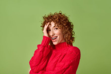 Young woman wearing a cozy red sweater playfully smiles with curly hair on green background.
