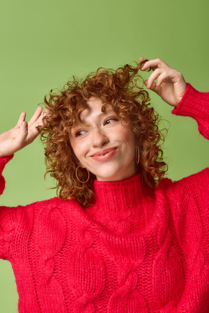 Bright and cheerful young woman with curly red hair playfully poses in a cozy red sweater.の写真素材