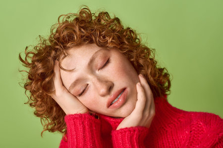 Blissful young woman with curly red hair rests her head gently on her hands, radiating warmth.