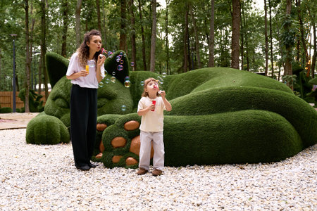 Mom and daughter share laughter while blowing bubbles near a playful green sculpture in a park.