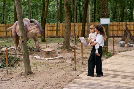 A mother holds her daughter close, admiring a life size dinosaur at an amusement park.の写真素材