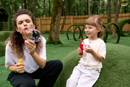 In a joyful amusement park, a mother and daughter share laughter as they create bubbles.