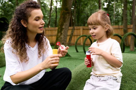 Smiling mother and daughter create bubbles together in a lush amusement park setting.