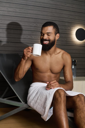 A shirtless man with a beard enjoys a peaceful moment while sipping a warm drink in a spa.の写真素材