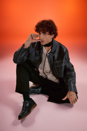 A stylish young man sits on the floor, showcasing his outfit and cool demeanor.