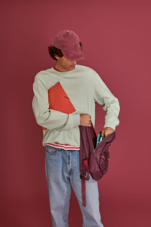 Fashionable man in casual clothing checks his backpack while holding a stylish notebook.