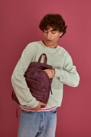 Handsome man wearing a light sweater reaches into his stylish backpack in a vivid setting.