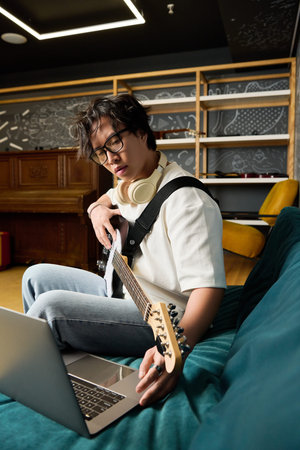 A young man in casual clothes strums his guitar in a stylish recording studio.