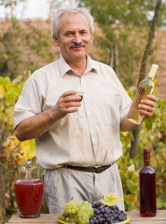 An elderly man holding a bottle and a glass of white vine in his hand, fresh grapes, a jug of must and a bottle of red vine in front of him on an old wooden table.の写真素材