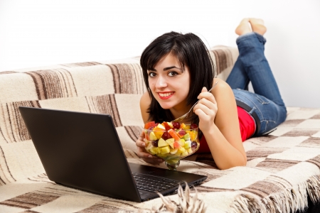 Smiling young woman laying in bed eating fruit salad in front of laptop.の写真素材