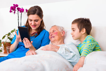 Elderly wiman in a nursing home with her grandson visiting, a doctor keeping them company, using digital tablet.の写真素材
