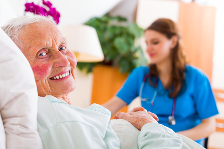 Happy senior woman laying in bed in a well equipped luxury nursing home with a caring nurse supervising her 24/7.の写真素材