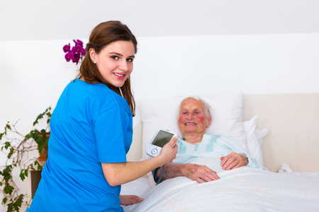 Happy nurse smiling to the camera while measuring senior woman's blood pressure in nursing home.の写真素材