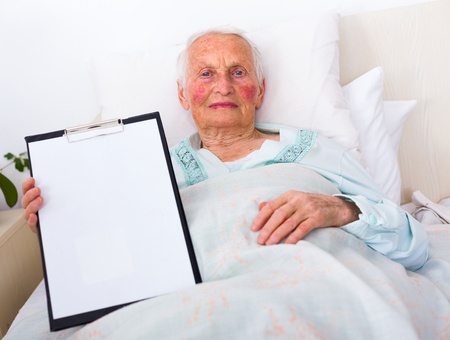 Elderly woman showong an empty paper on a clipboard , laying in bed in a nursing home.の写真素材