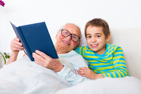 happy boy laying in bed with her grandmother who is reading a book with farytales.の写真素材