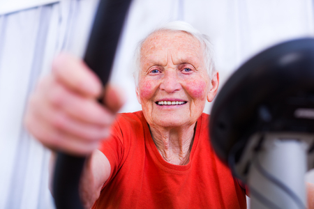Happy elderly woman spinning on indoor stationary bicycle in the nursing home.の写真素材