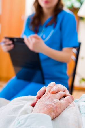 Senior patient's hands folded in bed in residential care home and a nurse in the background examining the patient.の写真素材