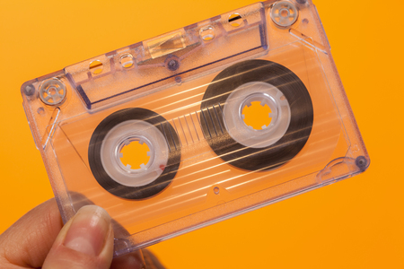 Hand holding transparent compact audio cassette tape - against orange backgroundの写真素材