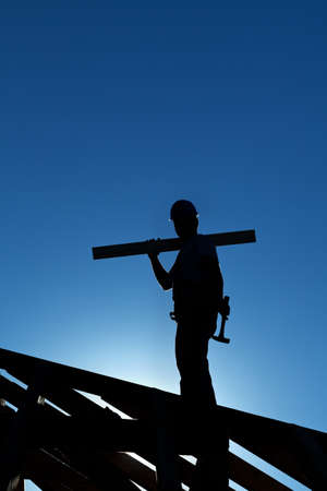 Worker silhouette on roof structure of building holding hammer and wooden plank backlit by the setting sunの写真素材