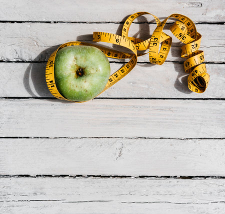 One green Apple wrapped in a flexible yellow centimeter tape lies on a white wooden table. Fitness...