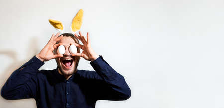 Charming Caucasian man with beard with yellow rabbit ears and two chicken eggs for eyes. Creative Easter bunny on white background, banner and plenty of space for your text or advertising.の写真素材