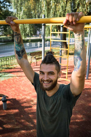 Young handsome Caucasian man pulls himself up on street sports field on warm summer morning. Strength training for muscles of arms. Active lifestyle concept. He stands and smiles.の写真素材