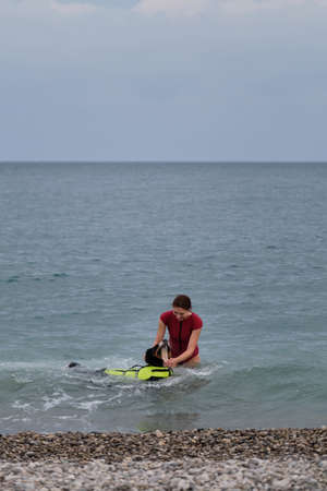 Young cute Caucasian red haired girl teaches her dog to swim in sea in special life jacket. Bernese Mountain Dog is lifeguard and bodyguard on water and on land. Training of rescue dog in pond.の写真素材