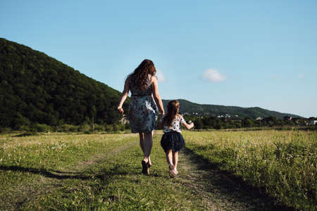 Young beautiful European mother in blue dress runs through chamomile field with her little daughter and laughs. Have fun with child in nature, the joy of motherhood. Rear view.の写真素材