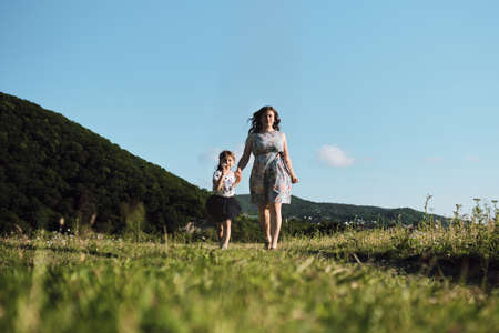 Young beautiful European mother in blue dress runs through chamomile field with her little daughter and laughs. Have fun with child in nature, the joy of motherhood. Front view.の写真素材