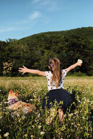 European girl on walk. Small Caucasian five year old girl with long wavy hair and polka dot dress is spinning and dancing in chamomile field, air bubbles are flying nearby and white pet dog is lying.の写真素材